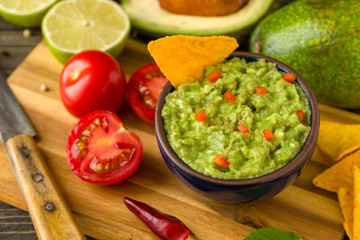 Guacamole in blue bowl on natural rustic desk with ingredient: lemon, tomatoes, peppers around.