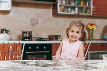 Little pretty kid girl in pink dress playing, smearing hands and face with flour in light kitchen at table. Child daughter cooking food, cookies in weekend morning at home. Happy childhood concept.