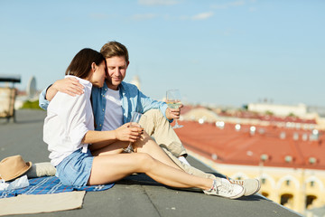 Amorous young man and woman with glasses of wine spending time on roof of building on sunny day