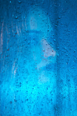 Art style portrait in blue tones of a lonely beautiful sad model girl behind a window glass, over which rain drops down