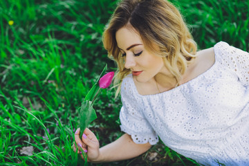 Young beautiful blonde girl in white dress with a flower tulip on a background of green grass