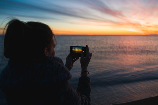 A silhouette of a young woman taking a photo of a sunset with her smartphone, making a memory.
