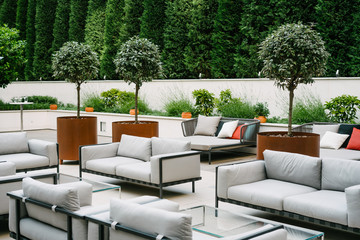 Beautiful, empty terrace with white sofas and glass tables, surrounded with green trees and...