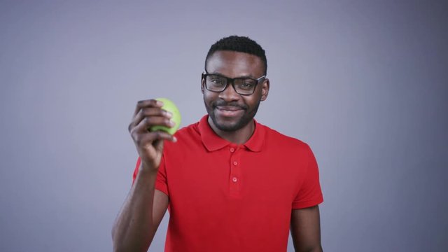 Handsome joyful young Afro-American man in nerdy like black spectacles juggling with apple, smiling straight to camera. Positive emotions, diet concept, healthy lifestyle. Male portrait on grey