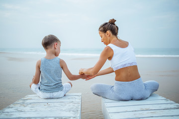 A mother and a son are doing yoga exercises at the seashore of tropic ocean