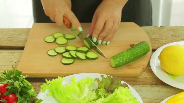 A female hand cuts cucumbers on a wooden board with a knife on a wooden board in circles. The camera moves from right to left. Near the table there are vegetables.