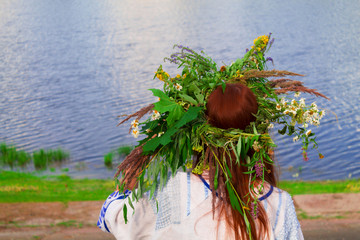 A girl in a national costume with a wreath of flowers on her head sits on the background of a river in summer