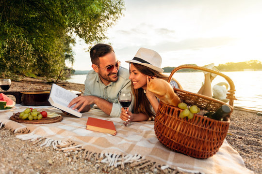 Happy young couple enjoying their time together at the beach. Lying on the blanket, reading books and drinking wine. Sunset over water in the background