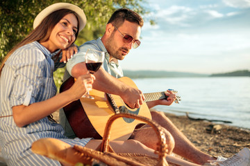 Obraz premium Happy young couple having a great time together at the beach. Man is playing guitar and singing, woman holding her head on his shoulder and enjoying. Sunset over water in the background.