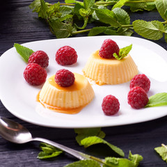Small portioned panna cotta with fresh raspberries. Milk creamy dessert, decorated with leaves of fragrant fresh mint. White dessert plate on a black wooden table.