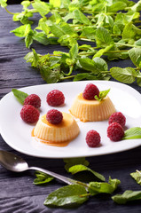 Small portioned panna cotta with fresh raspberries. Milk creamy dessert, decorated with leaves of fragrant fresh mint. White dessert plate on a black wooden table.
