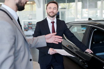 Smiling professional seller talking to his client while showing him a new car