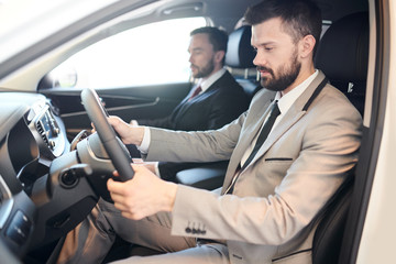 Bearded young man driving a new car with professional salesman sitting near by