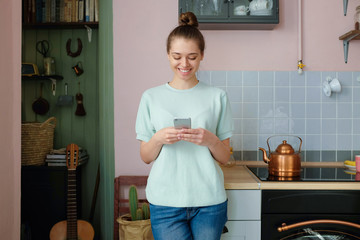 Horizontal portrait of young European woman standing alone on kitchen with smartphone in hands...