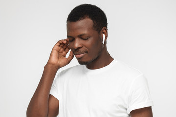 Closeup photo of young handsome African man turned a bit to left side of picture isolated on grey background holding wireless earphone with one hand listening to favorite tracks with closed eyes
