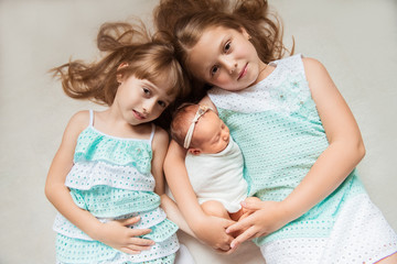 Lifestyle portrait of cute white Caucasian girls sisters holding little newborn baby, lying on bed blanket indoors. Family love bonding together concept