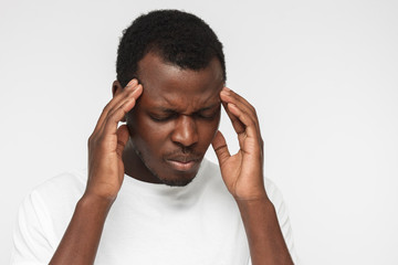 Indoor closeup of African American man pictured on gray background dressed in casual T-shirt suffering from terrible pain in head, showing it in facial expression looking helpless and miserable