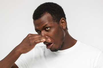 Horizontal indoor headshot of young African American man pictured isolated on gray background holding his nose because of unpleasant stinky odor he feels showing vivid face expression of disgust