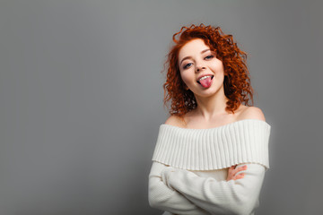 A young curly haired red-haired girl jokingly shows her tongue in the camera with her hands clasped...