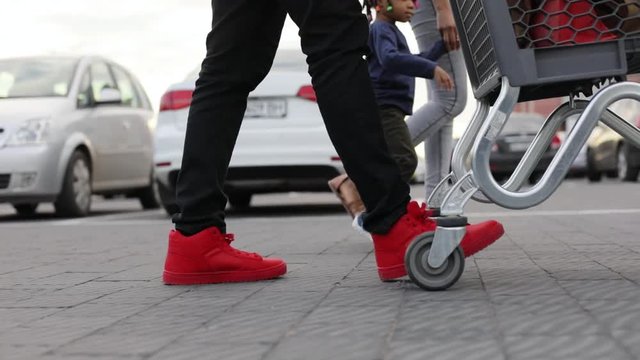 Low angle shot of a man in stylish red snickers pulling the shopping cart while walking between the parked cars, his wife and daughter walking near. Family portrait