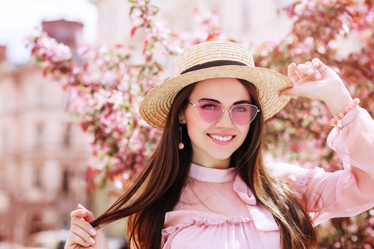 Outdoor close up portrait of young beautiful happy smiling woman wearing stylish pink cat eye sunglasses, straw hat, earrings, blouse, posing in street of city. Copy, empty space for text