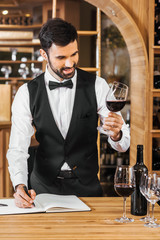 smiling young sommelier examining color of wine and making notes at wine store