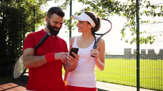 Male and female tennis opponents put off their tennis rackets, they use smartphone, the man shows something to smiling beautiful woman on his phone, she laughs at it. Having fun, relaxation, having a