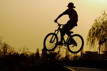Obraz premium Bike jumps/Silhouette of a jumping biker on a mountain bike in an outdoor park projected against the sunset orange sky. 