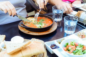 Family with child having traditional Israeli breakfast in the morning in cafe. Shakshuka, vegetable salad, lemonade, snacks - tuna, olives, cream cheese, thahin. Selective focus. Copy space.