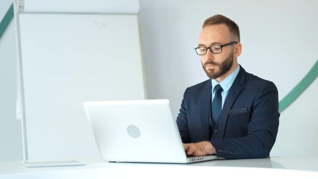 Attractive businessman in suit and glasses working with laptop in white office