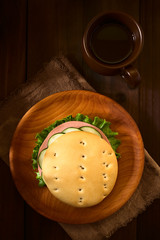 Traditional Chilean Hallulla bread roll prepared as a sandwich with lettuce, cold cut and cucumber served on wooden plate with a cup of tea, photographed overhead on dark wood with natural light