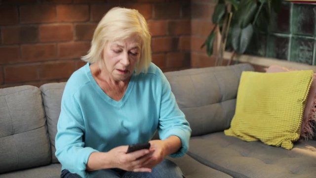 Caucasian old lady with backache touching pain place. Unhappy woman standing from gray sofa. Indoors. Lifestyle. Close-up.
