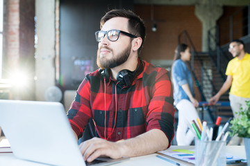 Portrait of modern bearded man wearing glasses and red shirt using laptop looking away pensively in...
