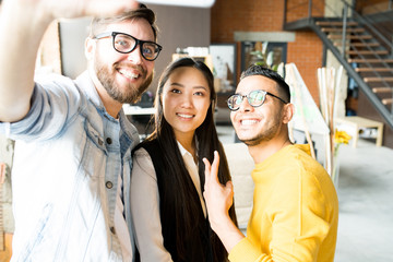 Multi-ethnic group of three creative young people smiling happily while taking selfie via smartphone standing in modern office, copy space