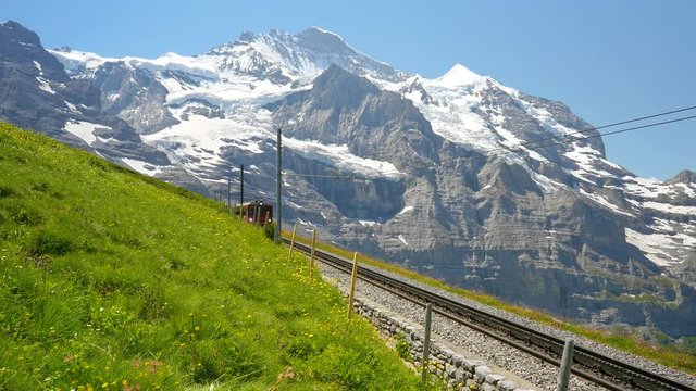 Static shot of a red train carrying tourists downhill from the Jungfrau Mountain range toward Kleine Scheidegg