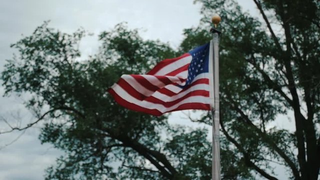 United States flag on a flag pole flapping in the wind in front of green trees with the image moving out of focus at the end
