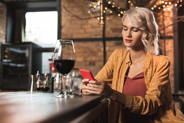 Pleasant pastime. Charming young woman sitting at the bar counter and surfing the Internet while drinking a glass of red wine