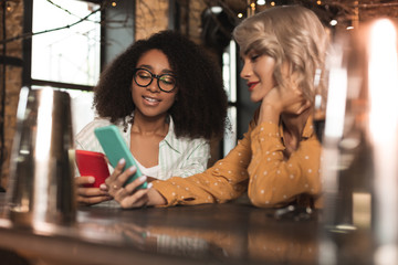 Pleasant interaction. Beautiful young women sitting at the bar counter and showing each other photos on their smartphones
