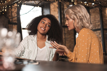 Full of skepticism. Pretty curly woman sitting at the bar counter with her friend and listening to...