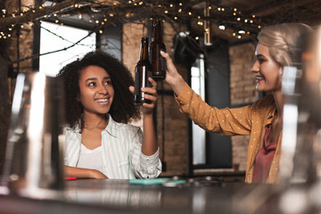 To us. Cheerful young women sitting at the bar counter and clinking their beer bottles while smiling