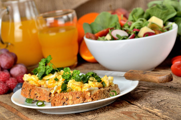 Wholemeal slice of bread with scrambled eggs, fresh herbs, spring onions, orange juice, tomatoes and salad bowl in background