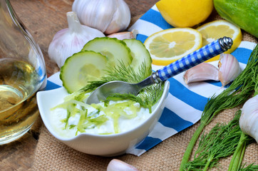 Spoon in bowl with Tzatziki - traditional Greek dressing or dip sauce, garlic, lemon, dill, cucumber, jug with oil and decoration in background
