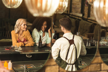 Pleasant evening. Charming young women sitting at the bar counter and communicating with a barman while drinking beer