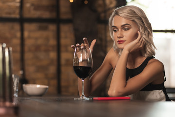 Deep in thoughts. Charming blonde woman running a finger along the rim of a glass of a red wine and thinking about something while sitting at the bar counter