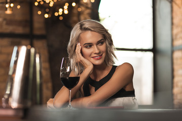 Welcoming smile. Charming blonde woman sitting at the bar counter, drinking wine and smiling at...
