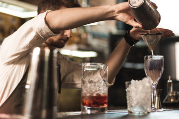 High professionalism. Young bristled barman making a cocktail while pouring it from the shaker into chinois thus straining the mixture