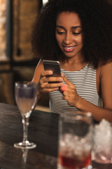 Good pastime. Beautiful curly woman sitting at the bar counter and texting her best friend while having a cocktail in bar