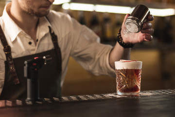 Last detail. Young professional barman sprinkling sugar over the glass with a cocktail, having mixed it by himself