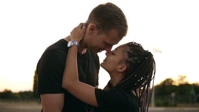 Side view of sensual young couple kissing while lying on car. Boy lay down her stylish girlfriend with dreadlocks on car hood. Outdoors