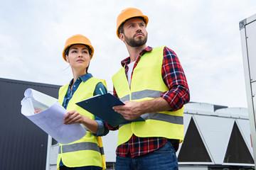 constructors in hardhats working with blueprints and clipboard on roof